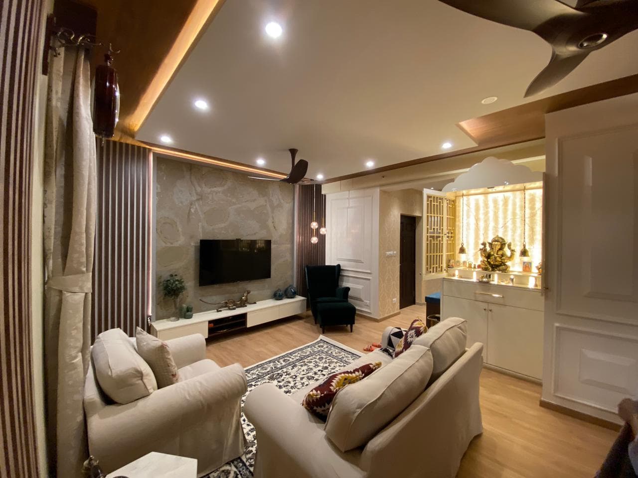 A warmly lit living room with stone-clad TV wall and pooja unit, designed by Anugraha Home Interiors